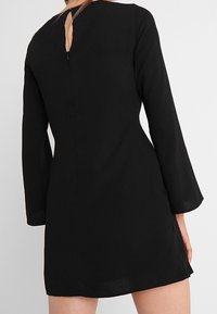 Svart klänning med långa klockärmar, en lätt utsvängning, detalj med nyckelhål på baksidan och ett mjukt, lätt tyg. Knälång nederkant.