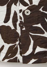 Gros plan sur un tissu floral marron et blanc avec un seul bouton blanc cousu près du centre.
