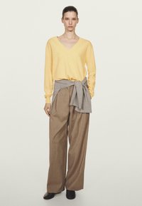 Gelbes V-Ausschnitt-Pullover, graue geschichtete Wickeljacke um die Taille, bequeme hellbraune weite Hose und schwarze Schuhe, vor einem neutralen Hintergrund.
