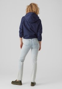 Marinblå hoodie-jacka med elastiska ärmslut, matchad med ljusblå jeans och gröna slip-on skor, sedd bakifrån. Släta texturer genomgående.