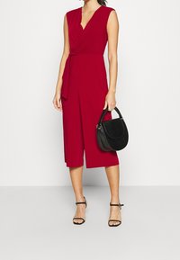 Combinaison sans manches rouge avec un lien à la taille et des jambes larges. Associée à un sac à main noir avec une finition texturée.