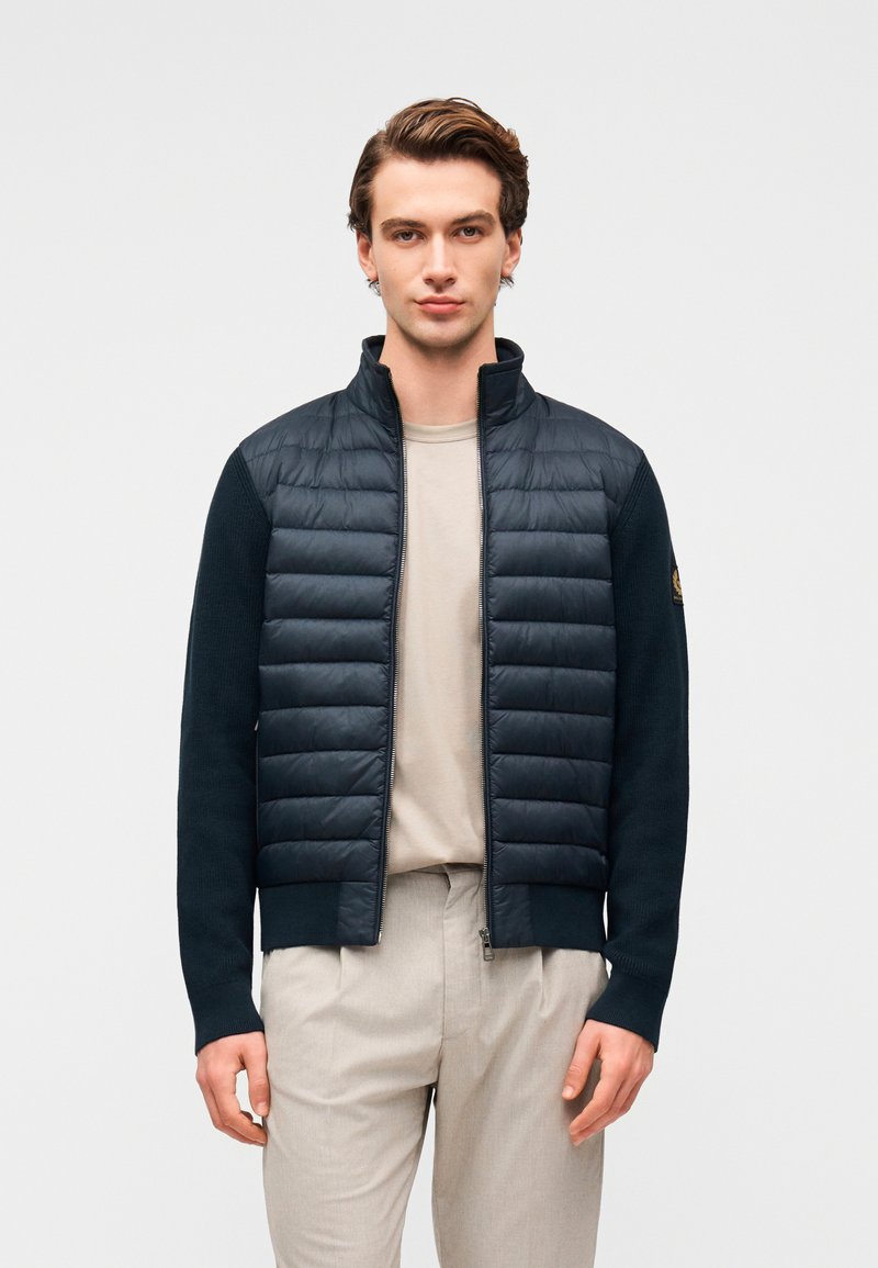 Σκούρο μπλε μπουφάν με καπιτονέ μπροστινό μέρος και μανίκια με ριμπ ύφασμα, διακοσμημένο με ψηλό γιακά και κλείσιμο με φερμουάρ, φορεμένο πάνω από μπεζ πουκάμισο και ανοιχτόχρωμο παντελόνι.