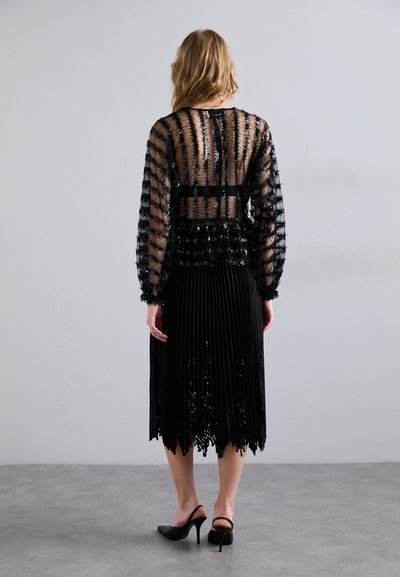 Fekete, réteges ruha áttetsző blúzzal, amely textúrázott részletekkel és plissé szoknyával rendelkezik, scallop szegéllyel végződik. Fekete magassarkú cipők.