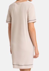 Donna che indossa un vestito beige chiaro alla ginocchia con maniche corte e dettagli di piping scuri sui polsini delle maniche e sull'orlo, vista da dietro.