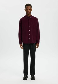 Bordeaux corduroy overhemd met knopen, lange mouwen, getailleerde pasvorm, gecombineerd met zwarte broek en zwarte schoenen op een effen achtergrond.