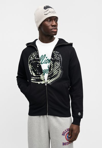 Μαύρο φούτερ με φερμουάρ και γραφικό σχέδιο, φορεμένο πάνω από ένα λευκό μπλουζάκι με λαμπερό κείμενο. Συνδυασμένο με γκρι φόρμα με λεπτομέρειες λογότυπου.