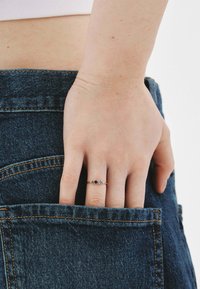 Anillo de oro con tres piedras preciosas, dos azules y una clara, colocado en una mano que se introduce en un bolsillo de unos vaqueros de mezclilla oscuros.