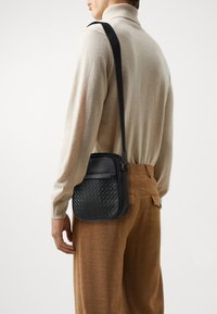 Black woven leather shoulder bag with smooth accents, rectangular shape, adjustable strap, and zipper closure, worn over a beige sweater.