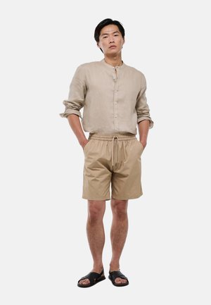 Homme debout, les mains dans les poches, vêtu d'une chemise beige boutonnée, d'un short beige à cordon et de sandales noires sur un fond uni.