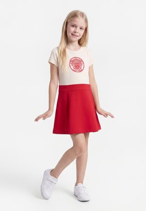 Fată tânără cu păr blond lung, purtând un tricou bej cu emblemă roșie și o fustă roșie, pozând cu mâinile ușor ridicate și zâmbind.