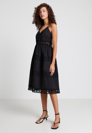 Donna con capelli ricci che indossa un vestito midi nero senza maniche con dettagli in pizzo e sandali neri con tacco e cinturino alla caviglia, in piedi di lato.