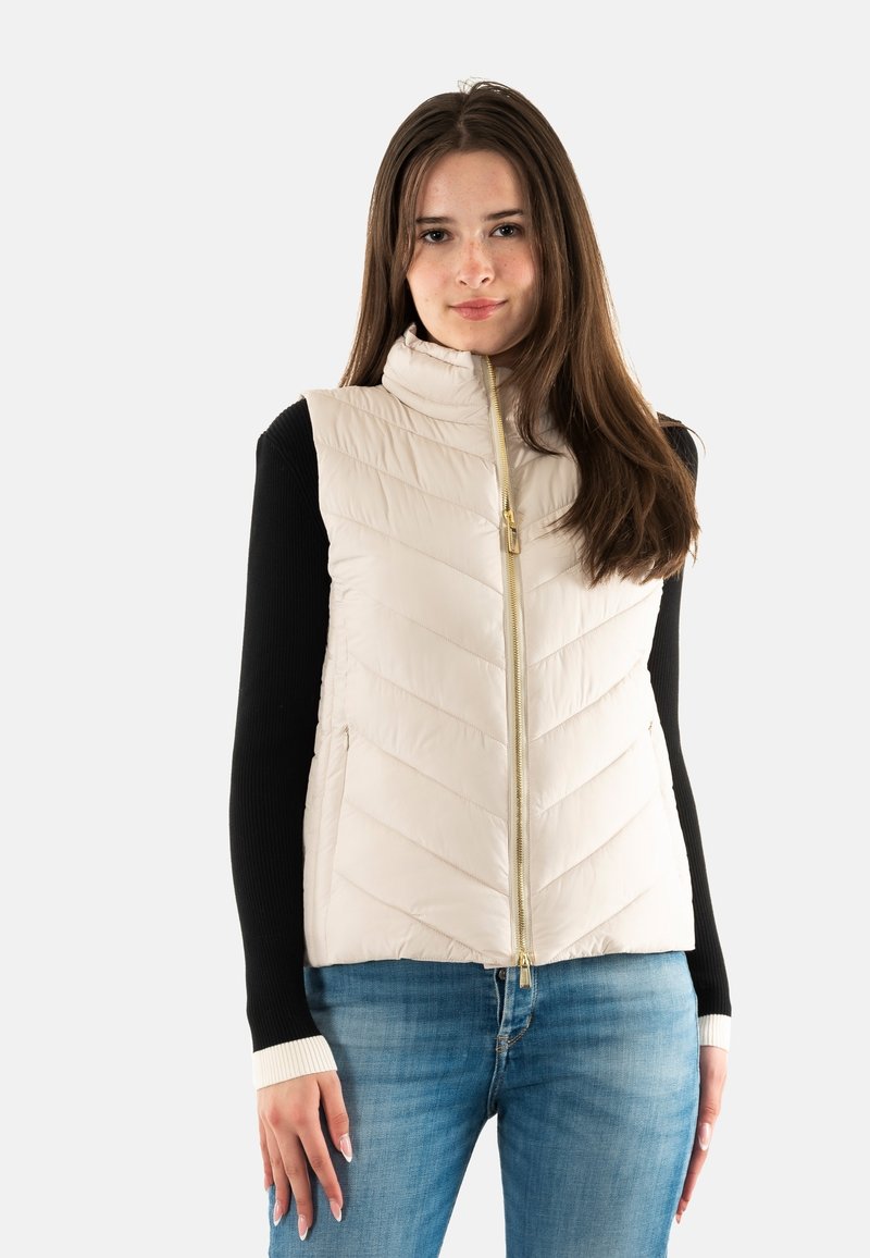 Gilet matelassé léger avec un col montant, un matelassage diagonal et une fermeture éclair dorée. Porté par-dessus un haut à manches longues noir avec des poignets blancs.