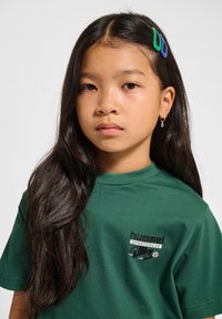 Grøn bomulds t-shirt med korte ærmer og rund halsudskæring, med "hummel SPORTSWEAR" branding på brystet. Barnet har langt sort hår med farverige clips.