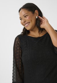 Top de encaje negro con un patrón floral texturizado, escote redondo y mangas tres cuartos. Pendientes de plata complementan el diseño.