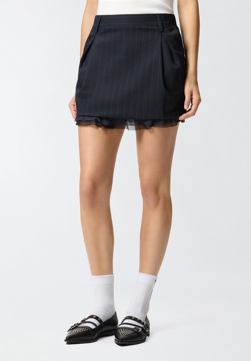 Tamna mornarsko plava mini suknja s nabranim detaljima, prugama, prozirnom volan-hemom i bočnim džepovima. Uparena s crnim cipelama i bijelim čarapama s prugama.