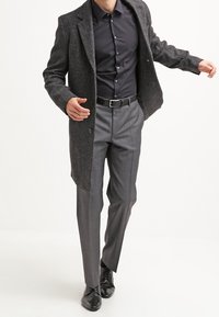 Tmavě šedý dlouhý kabát přes černou košili s knoflíky, doplněný šedými kostkovanými kalhotami a černými botami, zdůrazňující moderní a elegantní design.