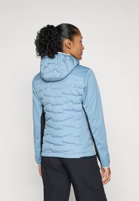 Vrouw met een krullende paardenstaart die een lichtblauwe gewatteerde jas met capuchon draagt, met de rug naar de camera gekeerd tegen een effen witte achtergrond.