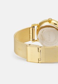 Montre en or avec un bracelet en maille. Boîtier rond avec un fond transparent. Le fermoir en métal présente le nom de la marque gravé, mettant en avant un design élégant.