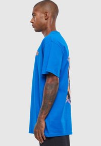 Lys blå bomulds t-shirt med korte ærmer, der har et grafisk print på ryggen. Sidesyn viser en afslappet pasform og rund halsudskæring.