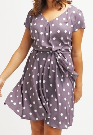 Woman wearing a short-sleeve lavender dress with white polka dots and a tied waist belt, standing against a plain background.