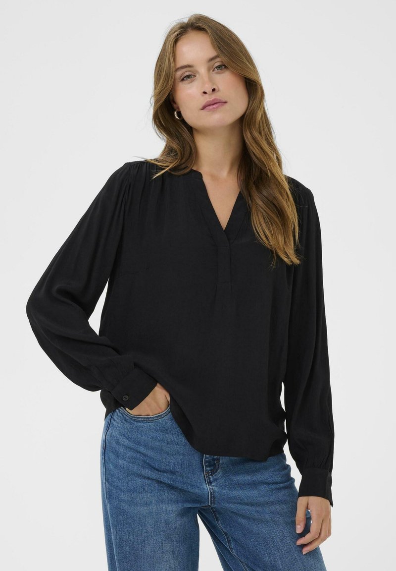 Sort bluse med V-udskæring, lange ærmer og rynke detaljer ved skuldrene. Parret med lyseblå jeans. Glat tekstur.