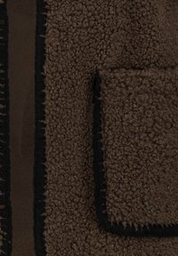 Braunes Fleece-Gewebe mit einer weichen, strukturierten Oberfläche. Schwarzer Besatz entlang der Kanten, mit einer Fronttasche mit strukturierter Oberfläche.