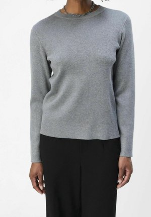 Personne portant un haut ajusté à manches longues gris clair et un pantalon noir, accessoirisée d'un collier chaîne argenté, debout devant un fond uni.