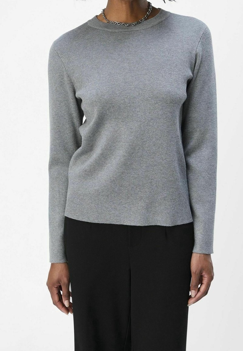 Personne portant un haut ajusté à manches longues gris clair et un pantalon noir, accessoirisée d'un collier chaîne argenté, debout devant un fond uni.