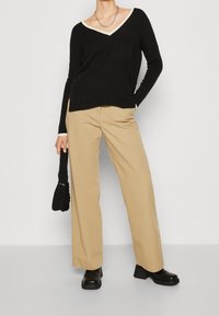 Maglione nero a maniche lunghe con colletto beige, pantaloni beige a gamba larga, stivali neri con tacco grosso e una borsa nera texturizzata.