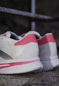 Hvide og lyserøde Adidas Adizero løbesko placeret på stentæppe med sløret mørk baggrund.