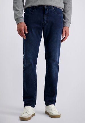 Man wearing dark blue jeans, gray sweater, and white sneakers standing against plain background.