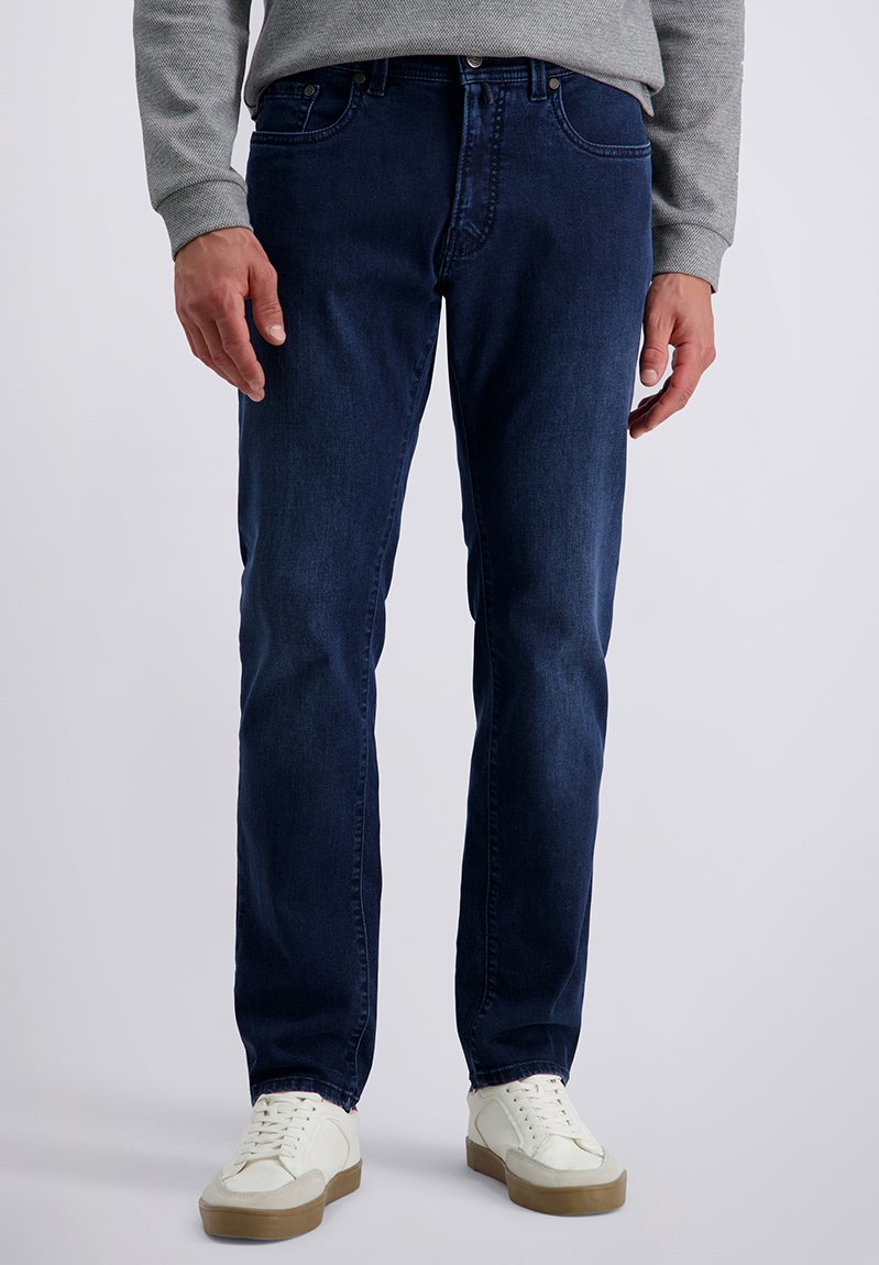 Man in donkere blauwe jeans, grijze trui en witte sneakers staand tegen een effen achtergrond.