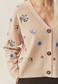 Hele roosa kardigan pehmest materjalist, kaunistatud värvikate lille- ja linnu tikanditega. Esiotsa kaunistavad silmatorkavad tumedad nupud.