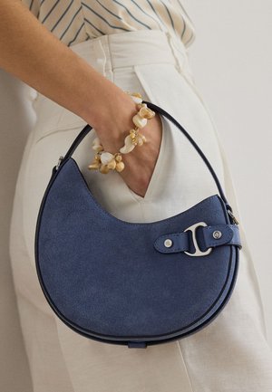 Το χέρι μιας γυναίκας στην τσέπη κρατάει μια τσάντα μισοφέγγαρο από μπλε σουέτ με ασημένια αγκράφα, φοράει εκρού βραχιόλι από κοχύλι και εκρού παντελόνι ψηλόμεσο.