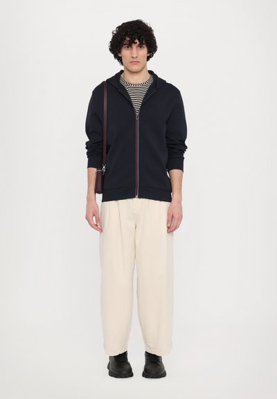 Sudadera navy con cremallera, debajo una camiseta de cuello redondo a rayas, pantalones de pierna ancha color crema y botas negras, complementadas con un bolso cruzado marrón.