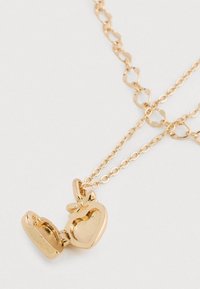 Collar de cadena dorada de doble capa con un colgante de medallón en forma de corazón abierto que presenta un pequeño lazo en la parte superior, colocado sobre un fondo blanco.