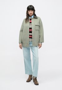 Ženska v svetlo zeleni preveliki jakni, črtastem pletenem puloverju, svetlo modrih opranih kavbojkah in rjavih gležnjarjih, stoji ob belem ozadju.