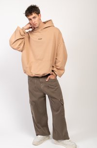Hanorac bej supradimensionat cu buzunar frontal și logo, asortat cu pantaloni cargo verzi oliv, lejeri, și adidași albi.