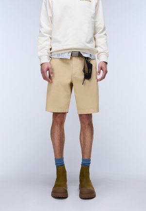 Mann trägt beige Shorts, weißen Sweatshirt, gestreiftes Hemd darunter, blaue Socken, olivgrüne Stiefel und eine schwarze Tasche, die an den Shorts befestigt ist.