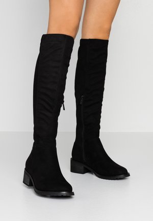 Schwarz wildlederne kniehohe Stiefel mit niedrigen Blockabsätzen und seitlichen Reißverschlüssen, getragen an nackten Beinen vor einem einfarbigen hellen Hintergrund.