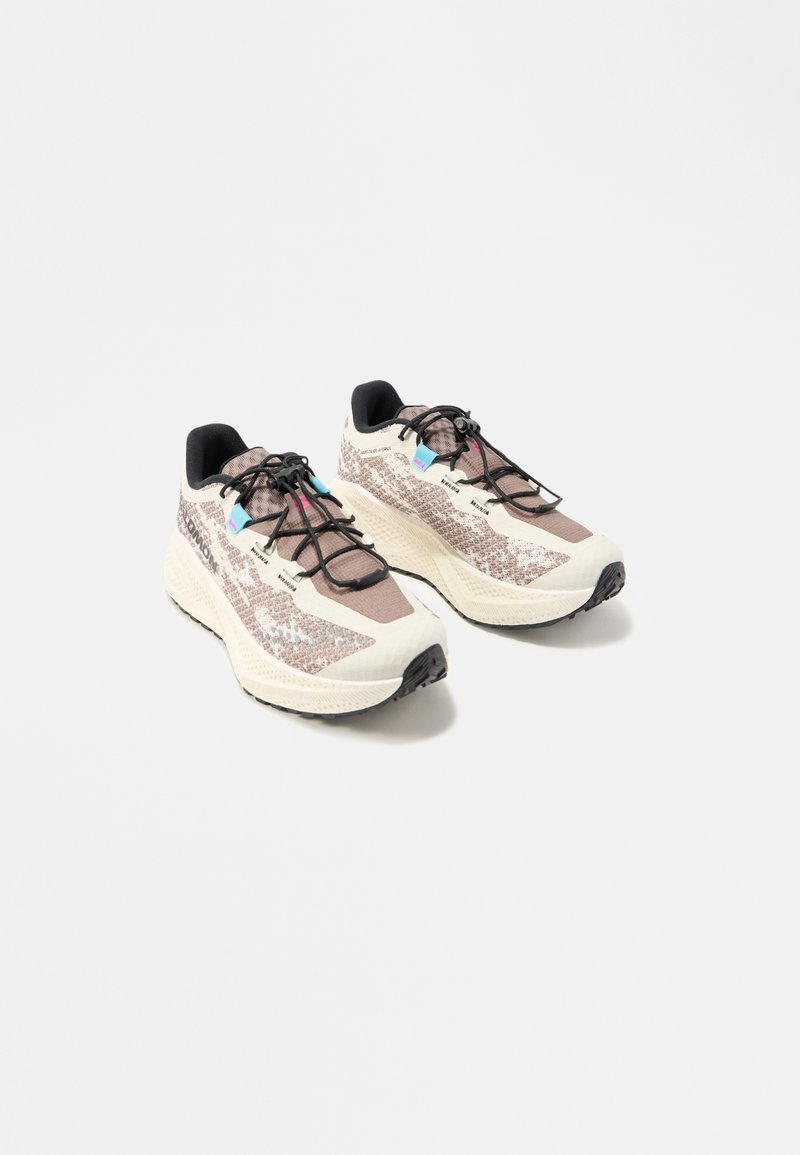 Paire de chaussures de course athlétiques beige et marron avec semelles noires, lacets noirs, touches de bleu et de rose, vues sur un fond blanc uni.