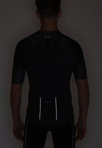 Musta urheilusuunnittelun vaatteet, joissa on lyhyet hihat, heijastava logo, selkäpaneelin muotoilu ja hienovaraisia ompeluyksityiskohtia.