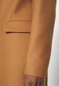 Bruine wollen jas met een soepele textuur, een op maat gemaakte pasvorm en een enkele borstzak. Klein merkembleem nabij de mouw.