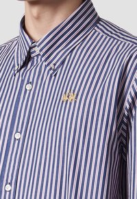 Camicia a righe in blu navy, con linee bianche e rosse, e un piccolo logo dorato vicino al petto. Tessuto testurizzato.
