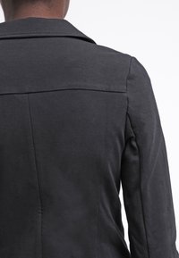 Černá bunda s hladkou texturou, strukturovaným límcem a švy vzadu, navržená s přiléhavým střihem a dlouhými rukávy.