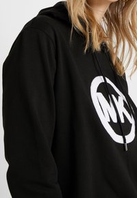 Felpa nera con cappuccio realizzata in tessuto morbido, con un grande logo circolare bianco sul davanti, cordini regolabili e una vestibilità comoda.