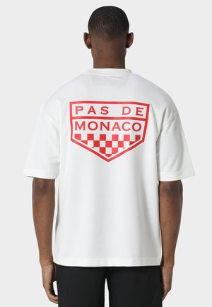 Homme portant un t-shirt blanc avec l'inscription rouge "PAS DE MONACO" et un motif à carreaux dans le dos, debout devant un fond clair uni.