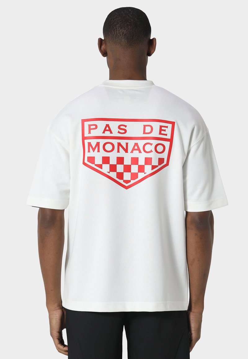 Άνδρας που φοράει λευκό T-shirt με κόκκινο "PAS DE MONACO" και σχεδιασμό καρό στην πλάτη, στέκεται σε απλό φωτεινό φόντο.