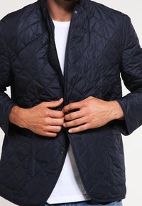 Marineblauer, gesteppten Jacke mit Stehkragen, Knopfverschluss und zwei vorderen Taschen. Der Stoff wirkt glatt mit einem leichten Glanz. Getragen über einem weißen Hemd.