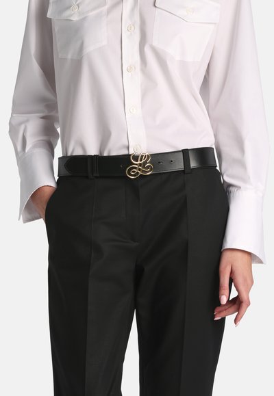 Persoon die een wit overhemd met knopen draagt en een zwarte broek met een zwarte riem met een gouden, verweven "L" gesp.